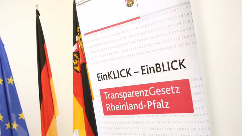 Roll-Up zum Transparenzgesetz Rheinland-Pfalz. Im Hintergrund drei Fahnen (Europa, Rheinland-Pfalz, Deutschland)